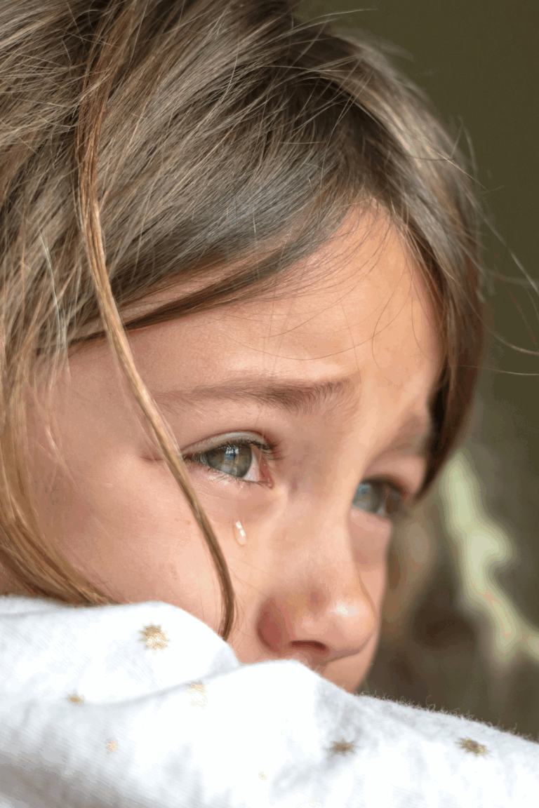 Sad crying Caucasian little girl with light brown hair and hazel eyes wearing a white, long-sleeved shirt with little flowers in front of a blurry, greyish brown background.